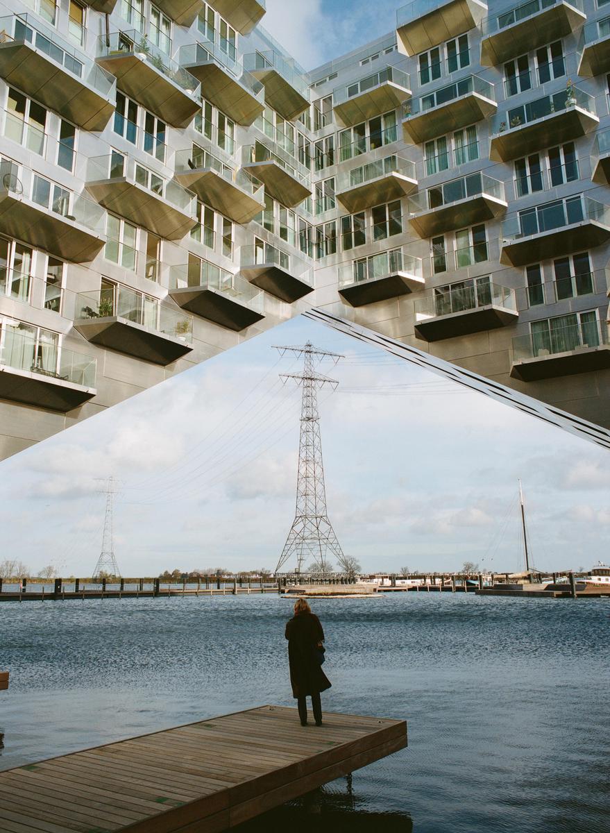A woman stands on a wooden dock looking out at the water with a power line tower in the distance and a modern building above.