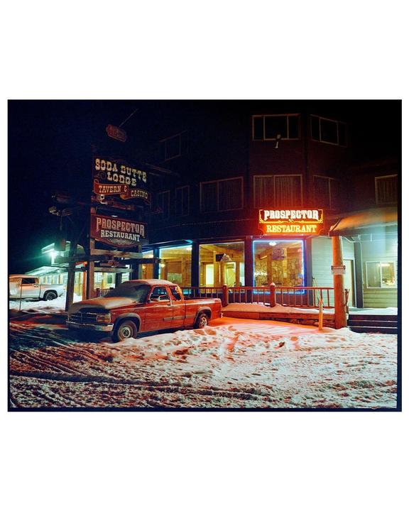 A red pickup truck is parked in the snow outside a lodge and restaurant at night.