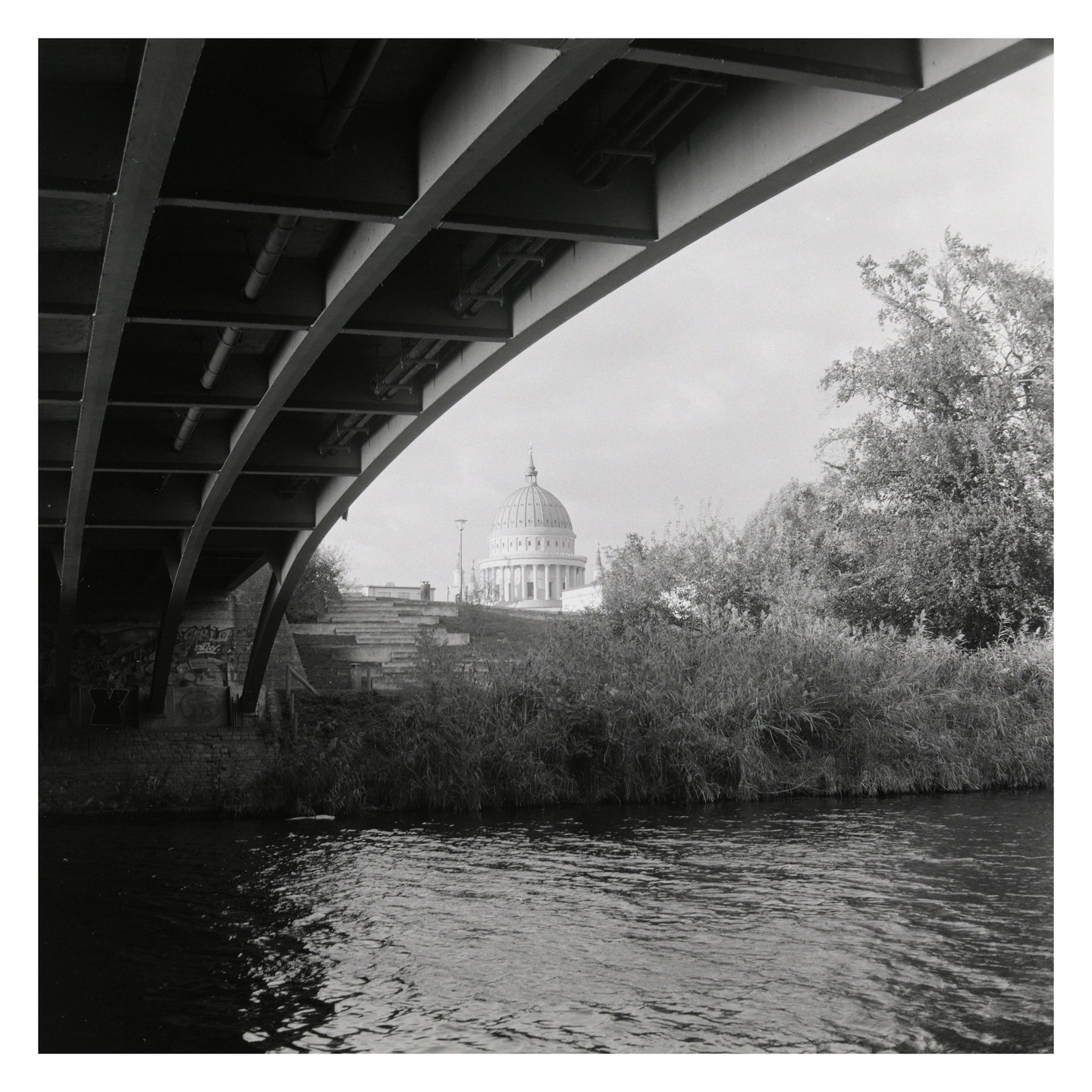 A domed building is visible from under a bridge over a body of water, with trees and bushes on the bank.