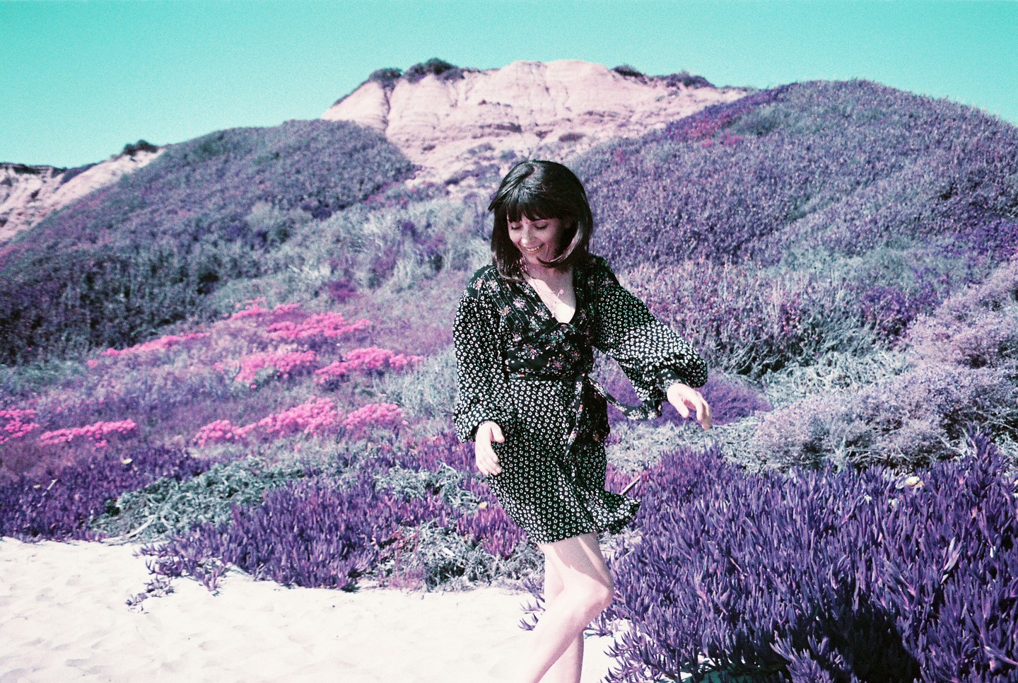 A woman in a patterned dress stands on a sandy path with purple and pink foliage and a large hill in the background.