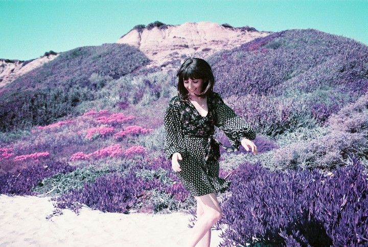 A woman in a patterned dress stands on a sandy path with purple and pink foliage and a large hill in the background.