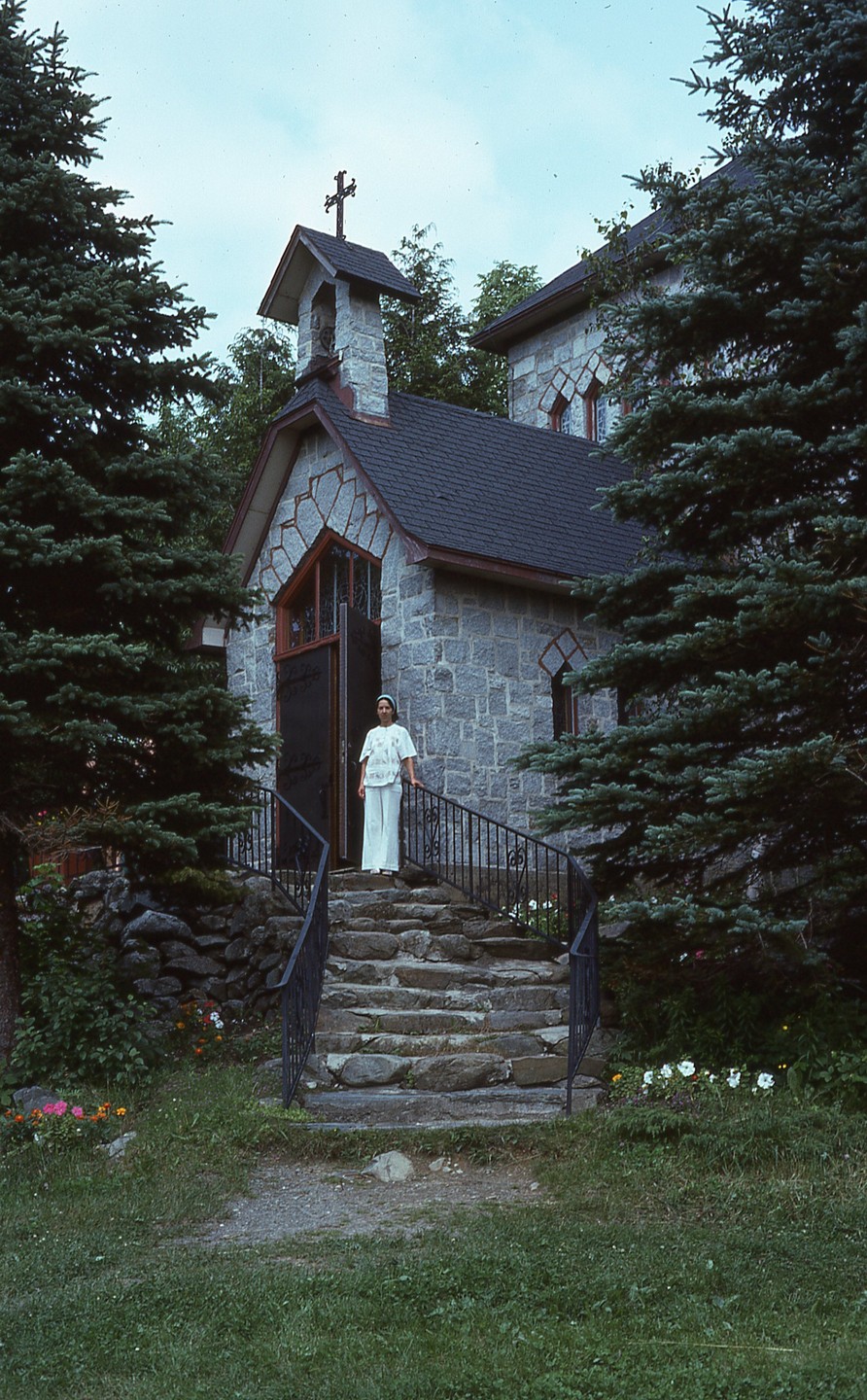 A woman stands on stone steps leading to a stone building with a bell tower and cross.