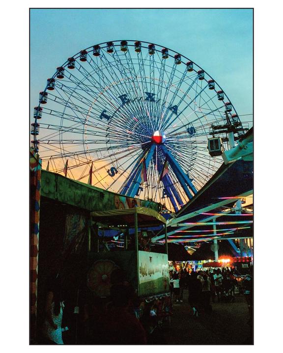 A large Ferris wheel with the word "TEXAS" spelled out in lights dominates the sky above a bustling fairground at dusk.
