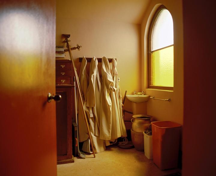 A room with robes hanging on a rack, a sink, and an arched window with yellow glass is seen through an open door.