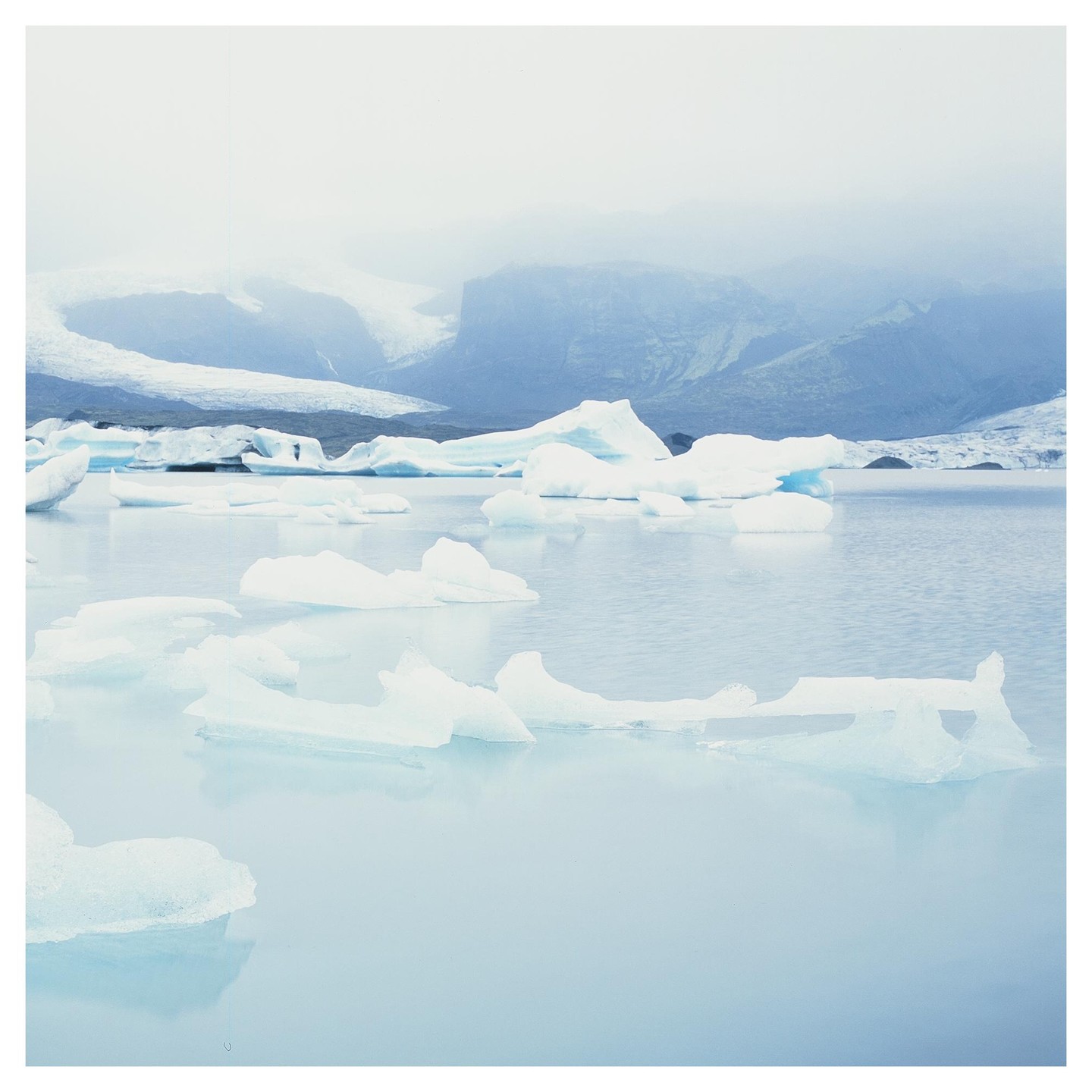 Icebergs float on a glacial lagoon with mountains and a glacier in the background under a hazy sky.