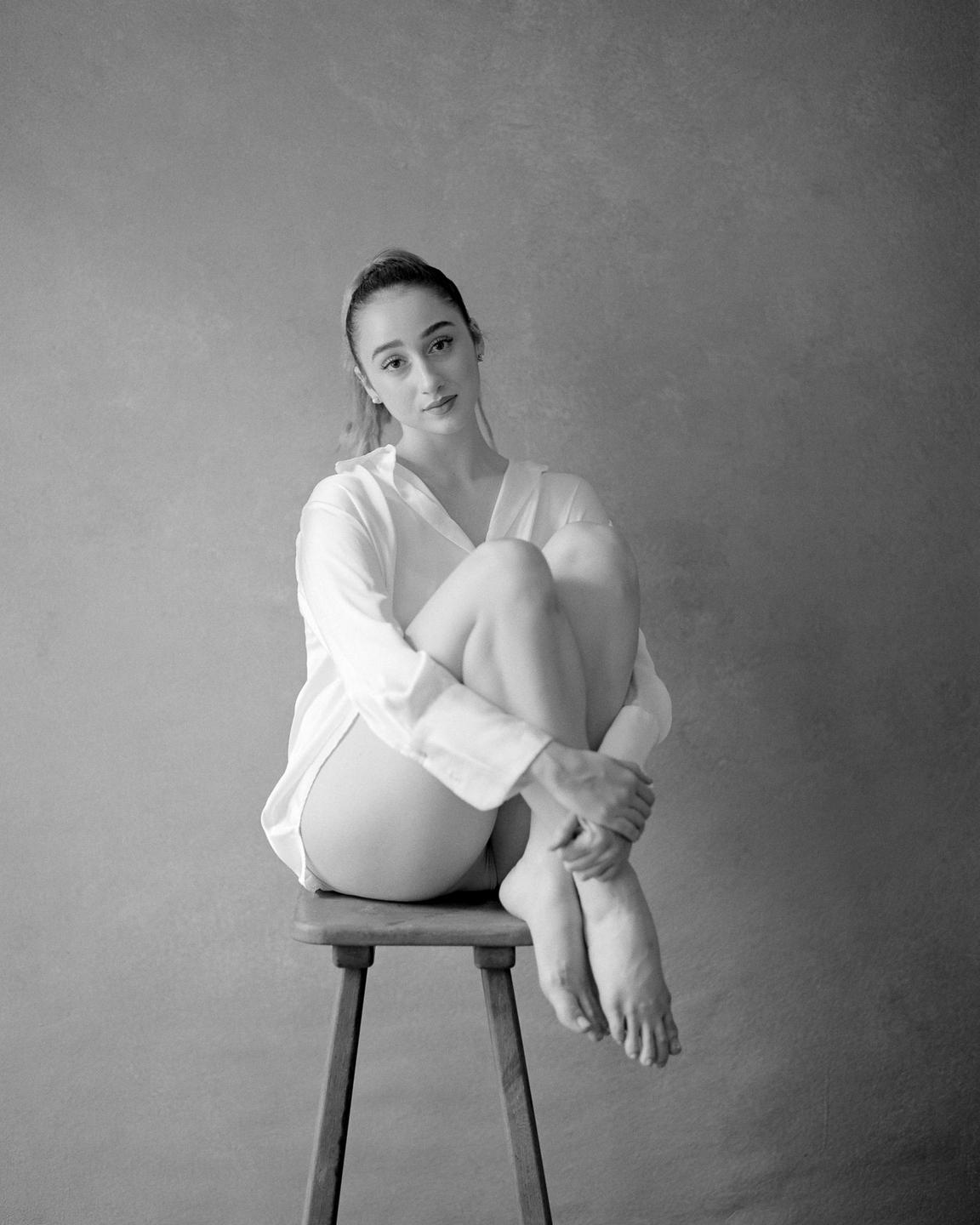 A woman wearing a white shirt sits on a stool with her knees pulled to her chest.