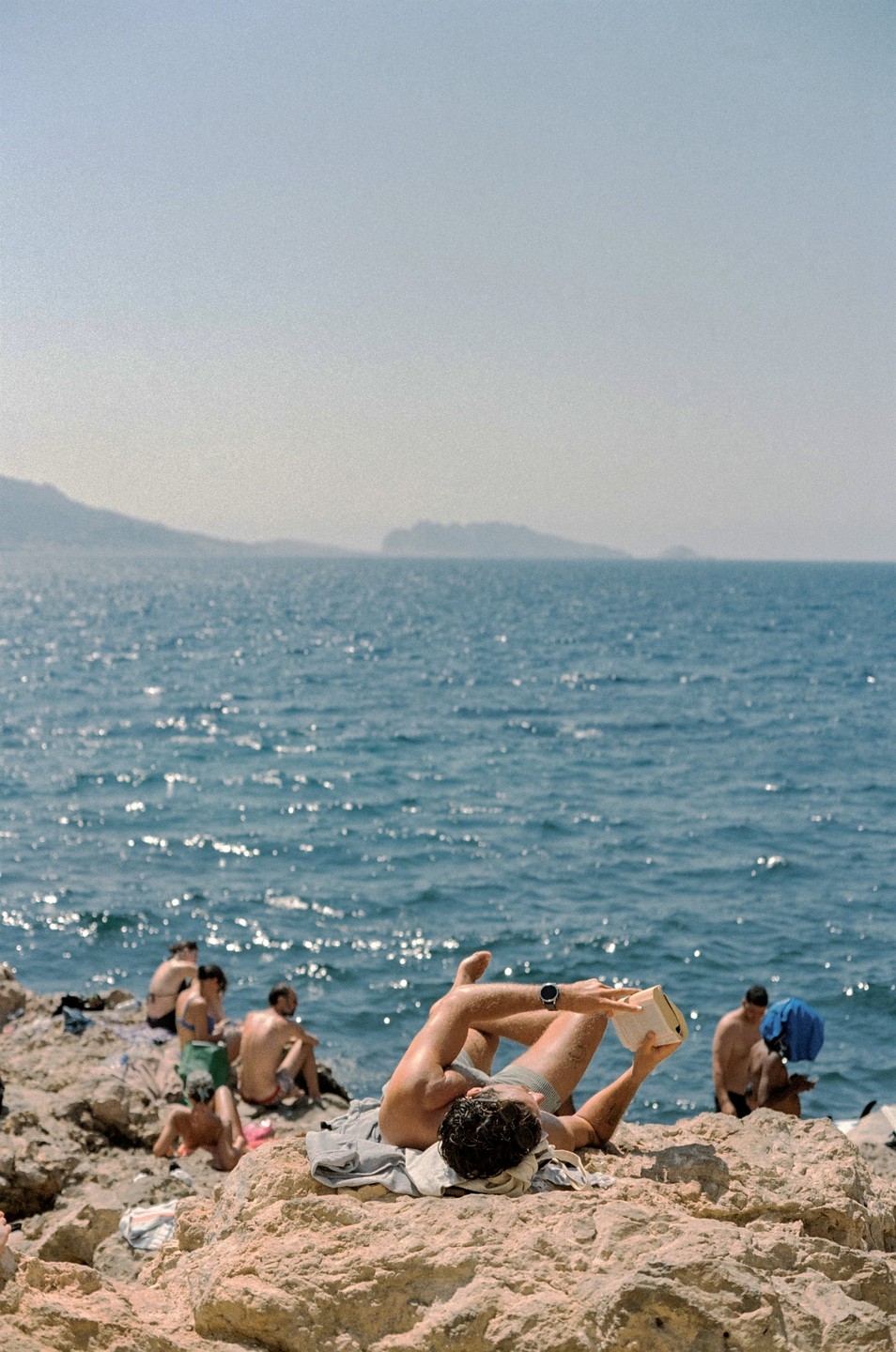 A man lies on his back on a rocky beach, reading a book, with other people and the sea in the background.