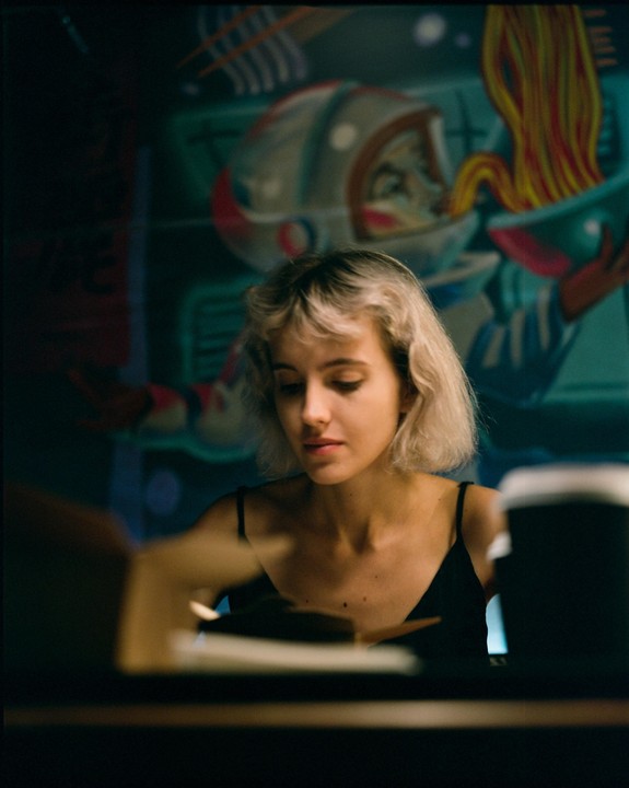 A woman with short blonde hair looks down at a table in front of a mural of an astronaut eating noodles.