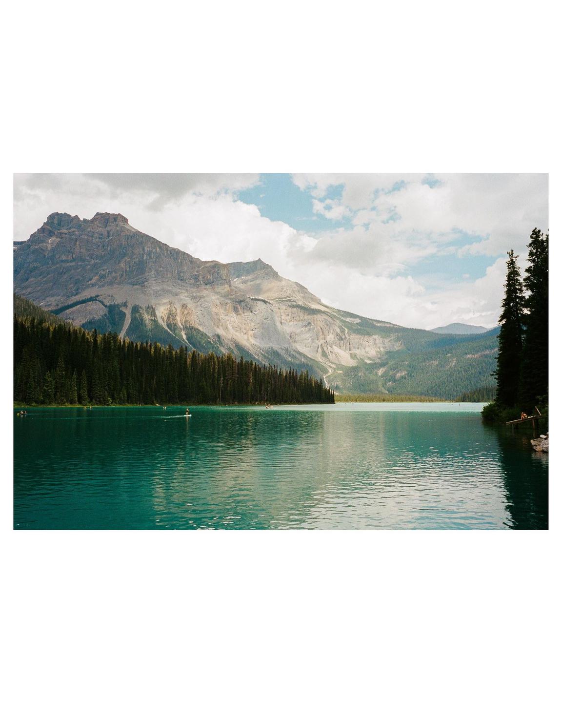 A turquoise lake is surrounded by a dense forest and a large mountain under a cloudy sky.