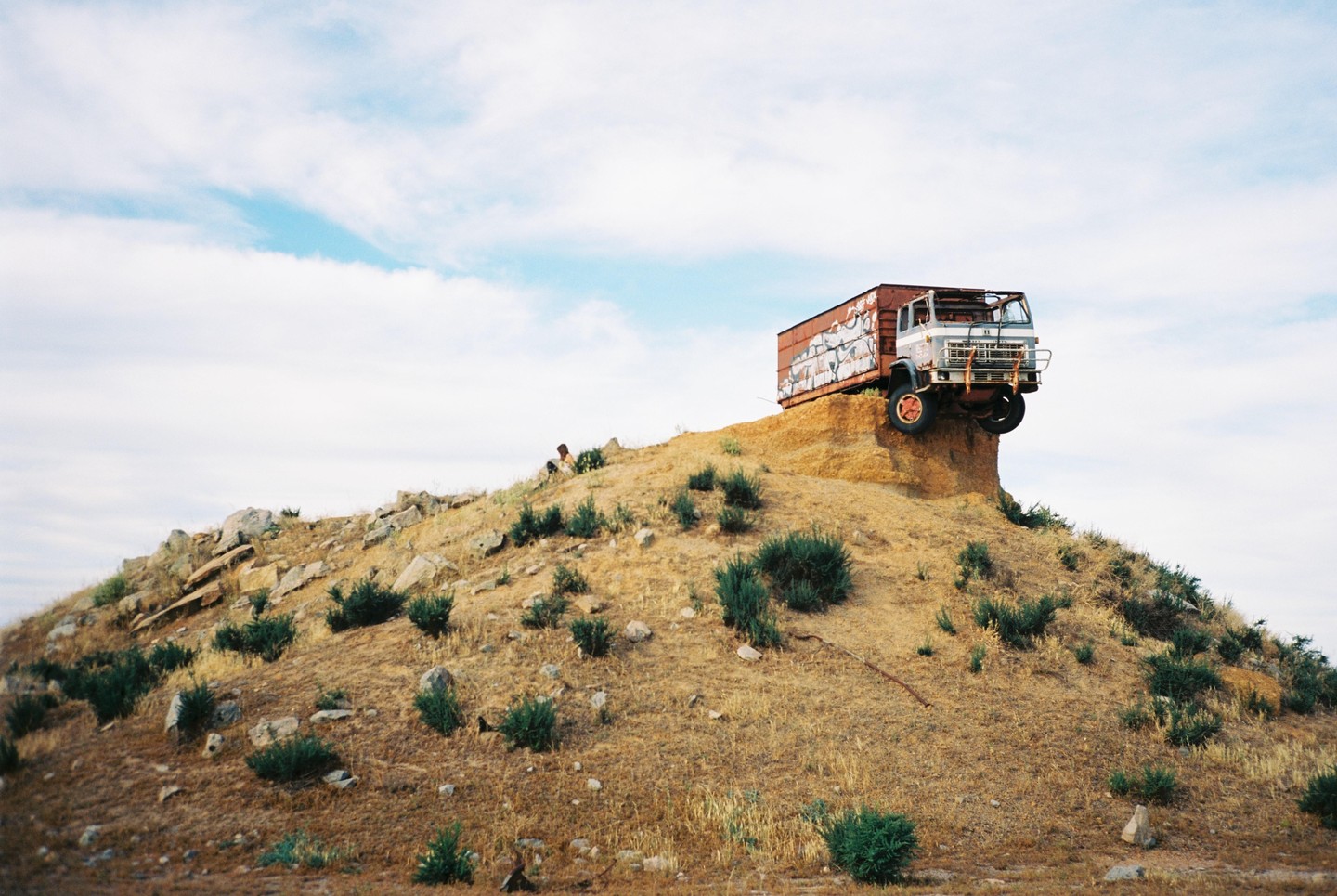 A truck is stuck on a hill with a person sitting nearby under a cloudy sky.