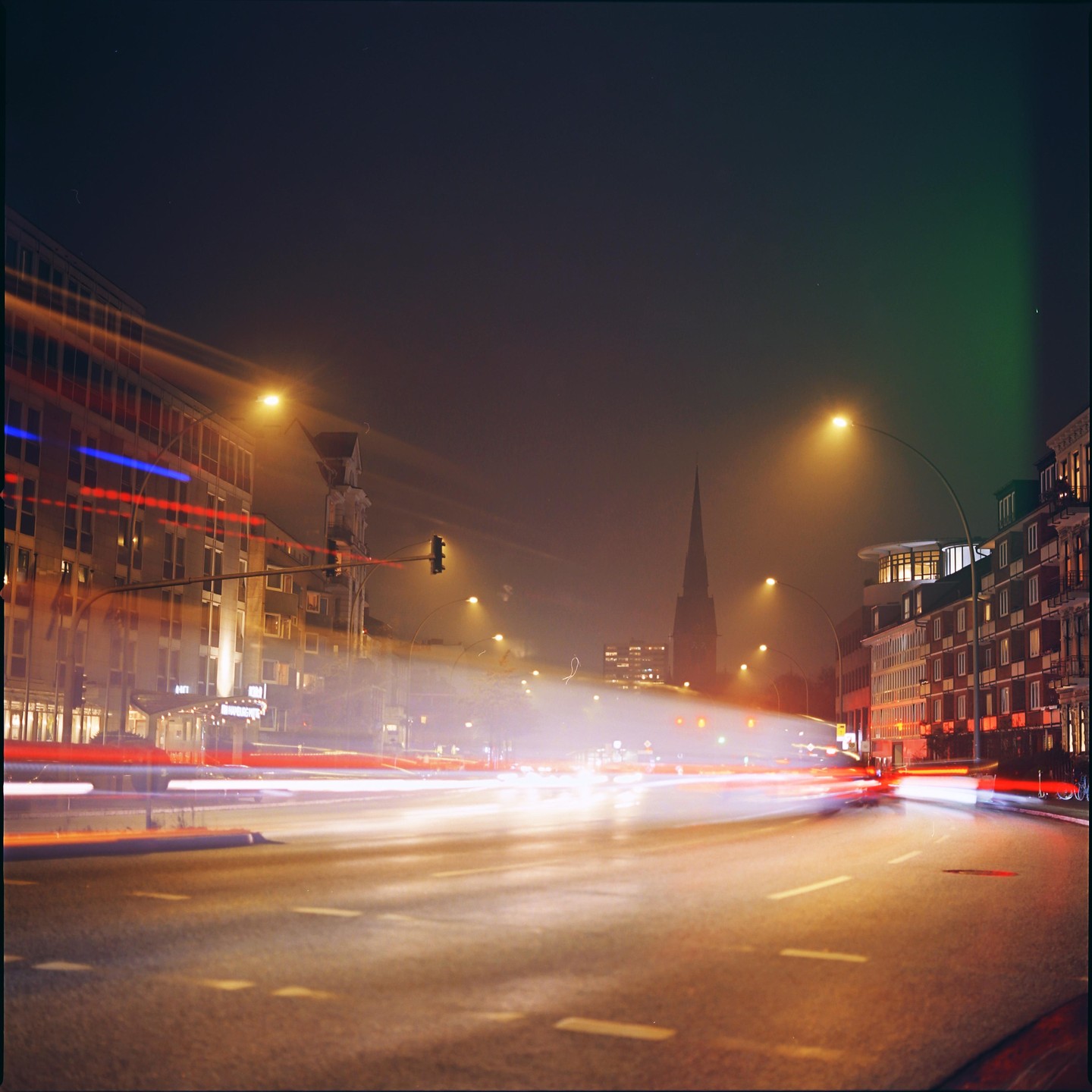 A long exposure shot of a city street at night shows light trails from vehicles and a church steeple in the distance.