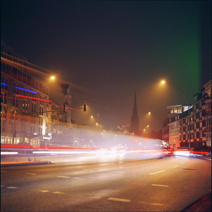 A long exposure shot of a city street at night shows light trails from vehicles and a church steeple in the distance.