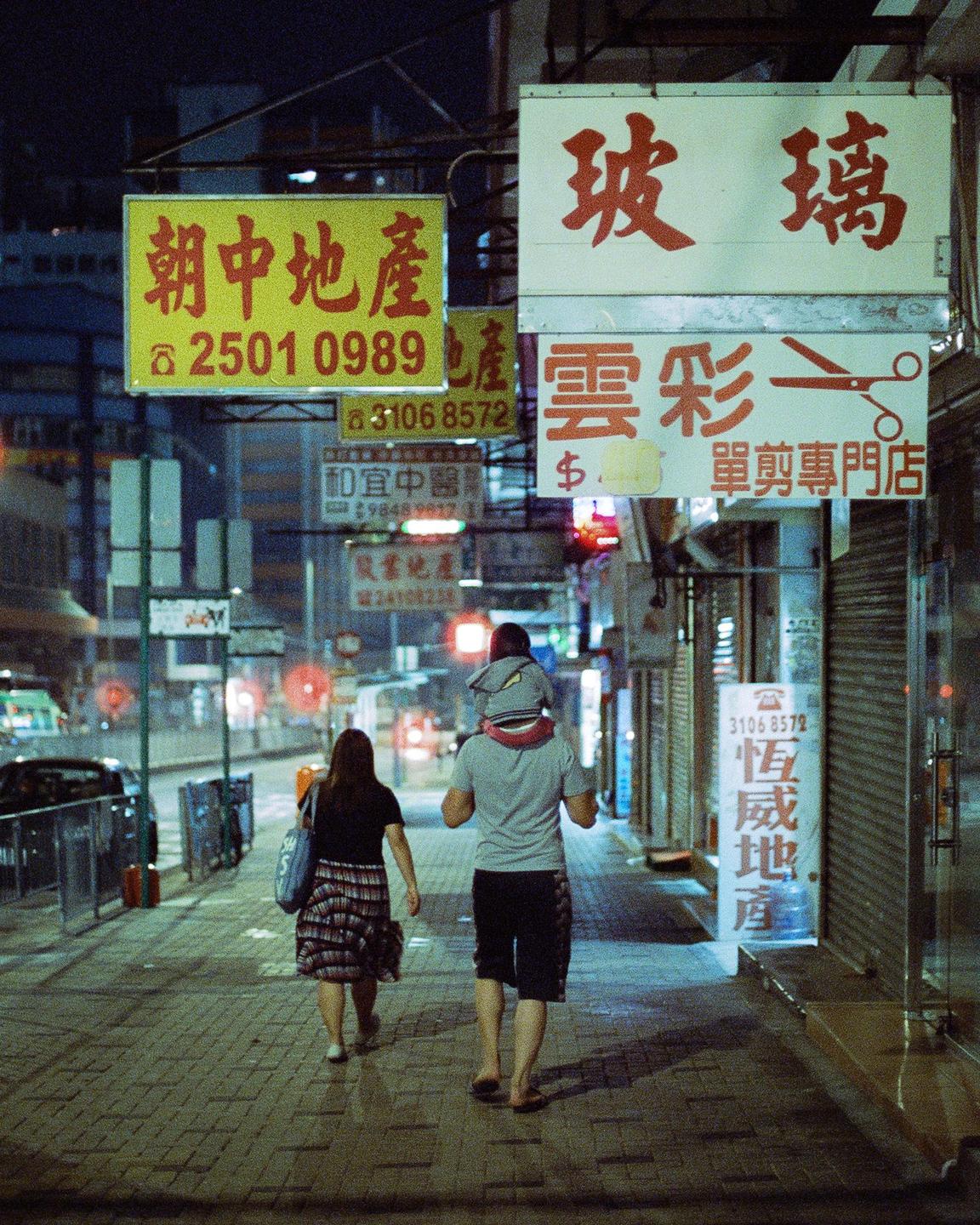A family walks down a city street at night, passing storefronts with neon signs.