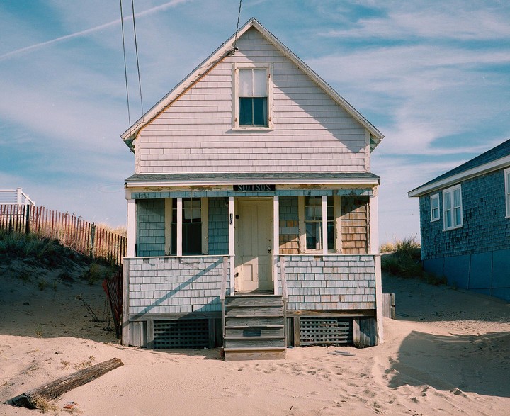 A small cottage with a porch and steps is surrounded by sand dunes under a blue sky.