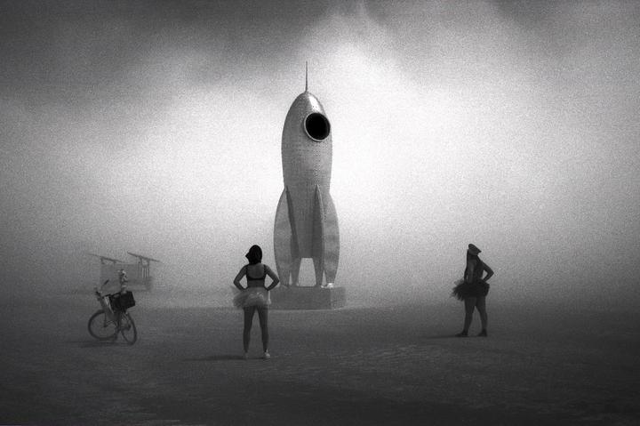 Two women stand in a hazy, open space with a large rocket sculpture and a bicycle.
