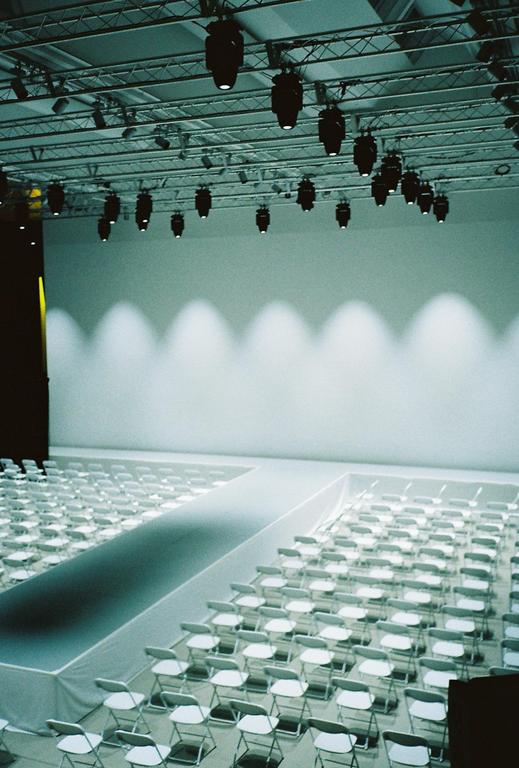 A fashion show runway is set up with rows of empty white chairs and spotlights illuminating a white wall.