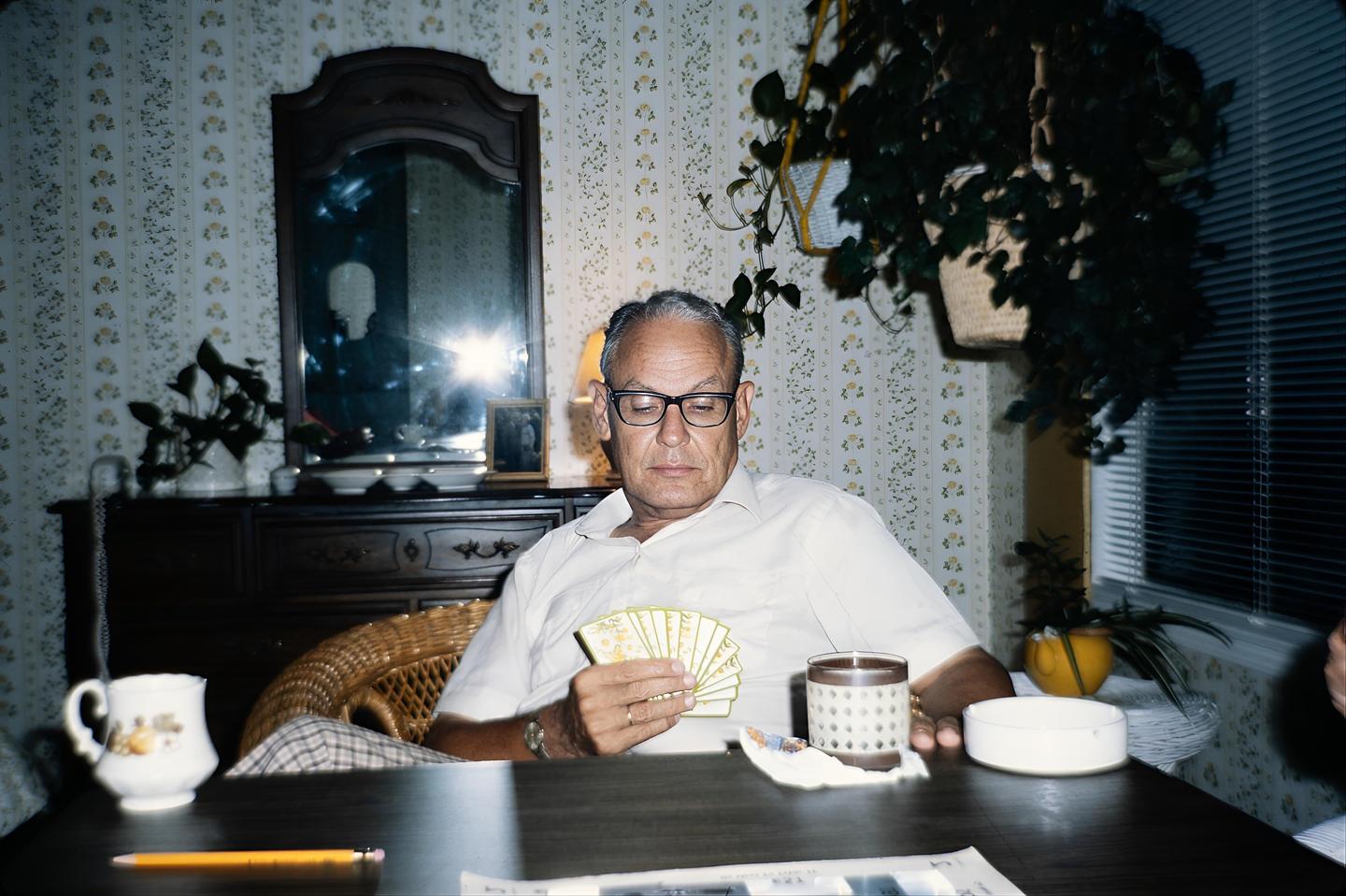 A man wearing glasses sits at a table and holds a hand of cards.