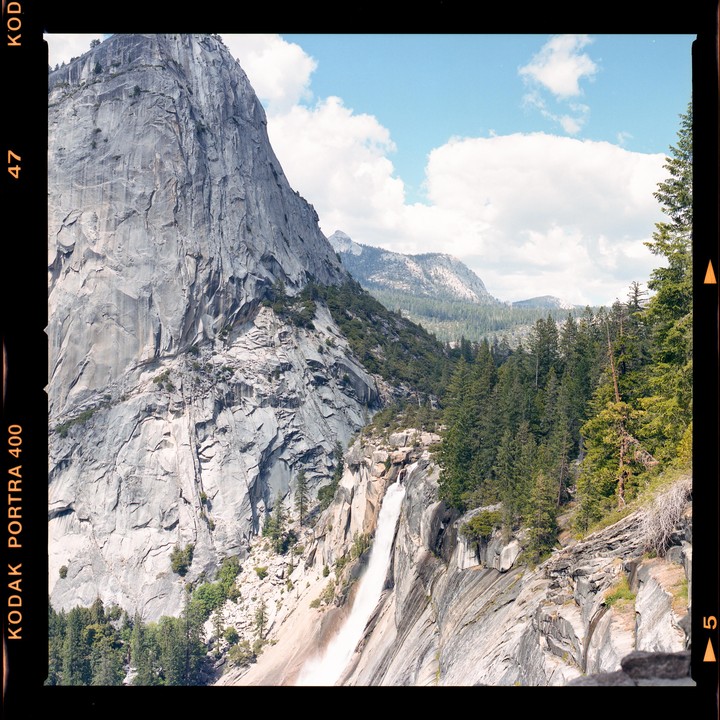 A waterfall cascades down a large granite mountain face next to a forest under a blue sky with clouds.