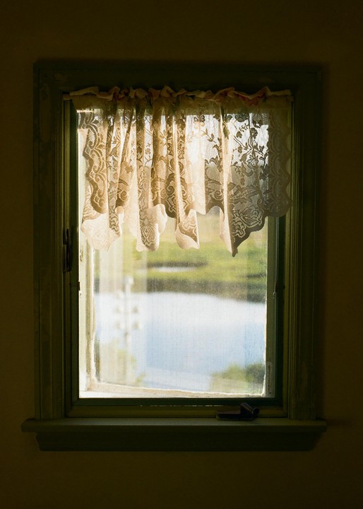 A window with a lace curtain looks out onto a body of water and green landscape.