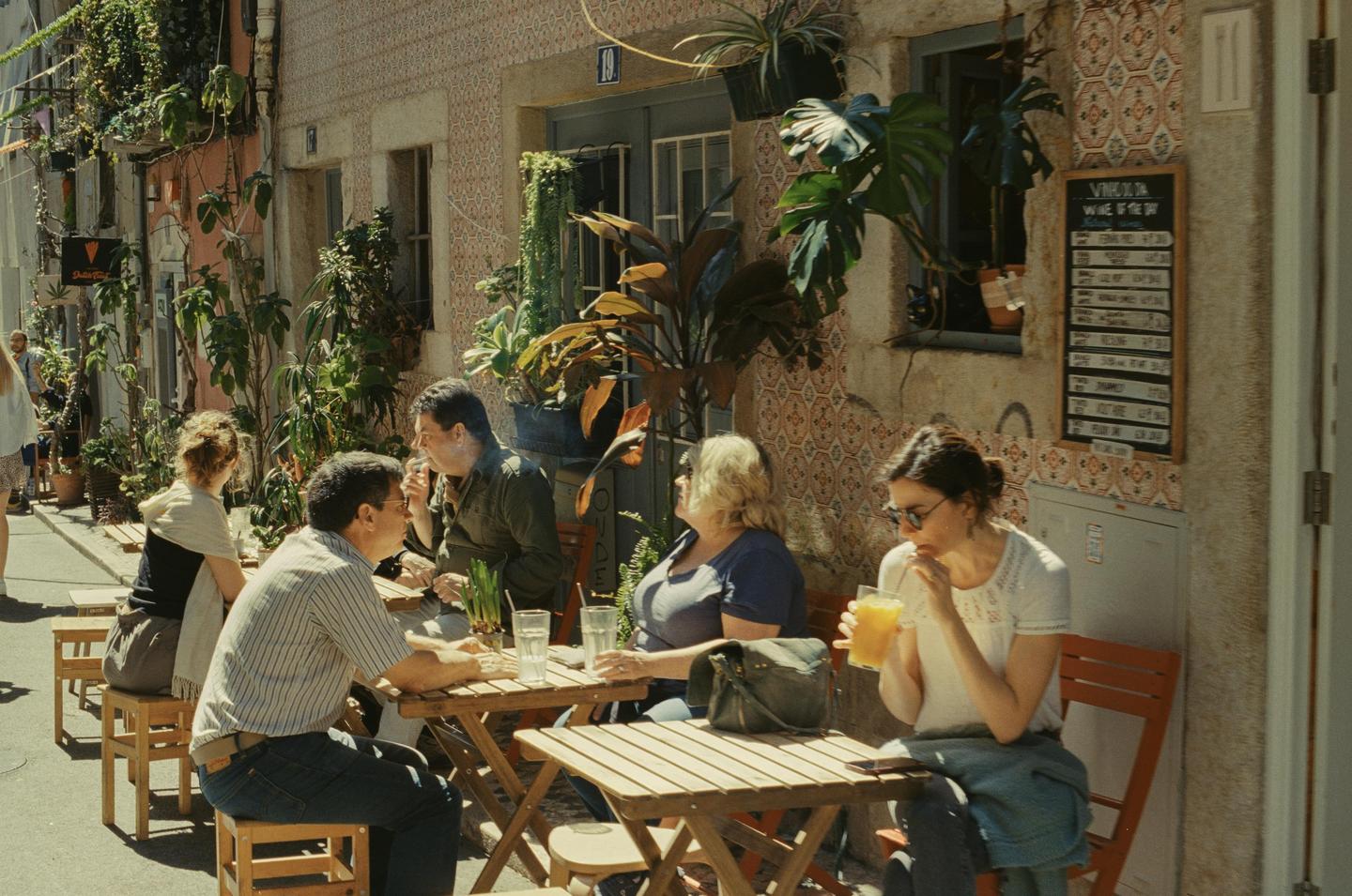 People sit at wooden tables outside a cafe on a sunny street lined with potted plants and vines.