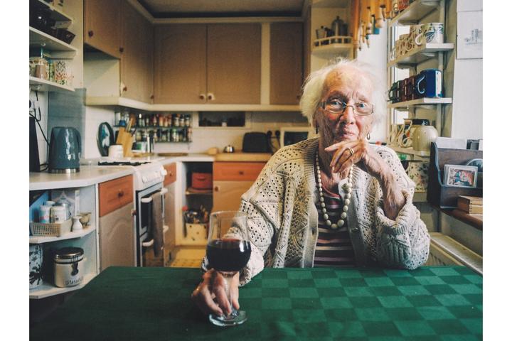 An elderly woman with white hair and glasses sits at a green checkered table in a kitchen, holding a glass of red wine.