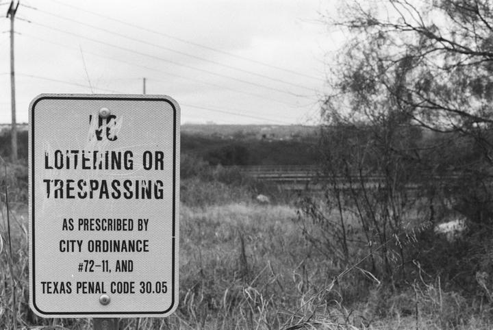 A "No Loitering or Trespassing" sign stands in a field with trees and power lines in the background.