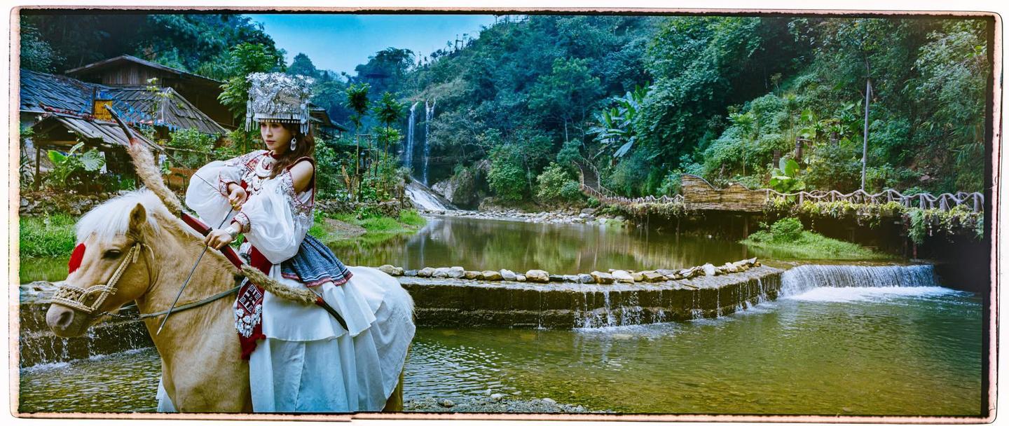 A woman in traditional attire with a bow and arrow sits on a horse in a lush, green landscape with a waterfall and river.