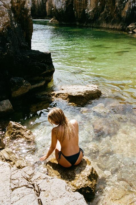A woman sits on a rock at the edge of a clear, green body of water surrounded by rocky cliffs.