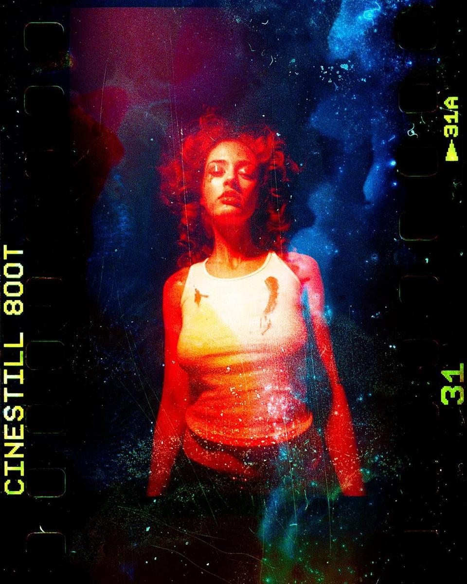 A woman with closed eyes and red hair is illuminated by red and blue light against a dark, textured background.
