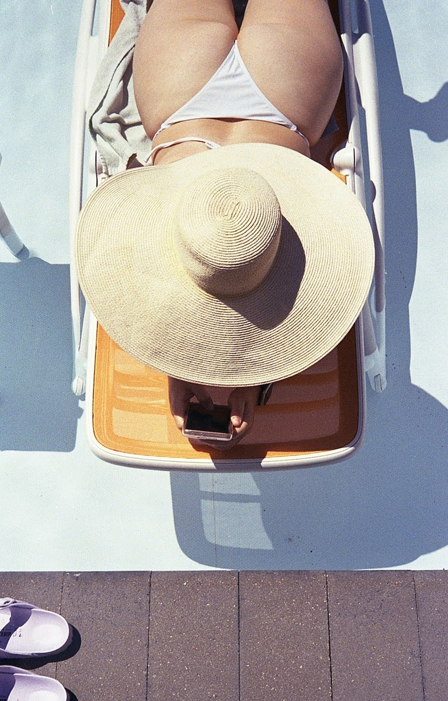 A woman in a white bikini and a large straw hat lies on a lounge chair by a pool, holding a phone.