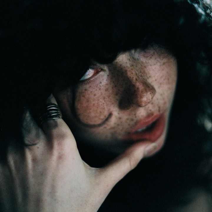 A woman with freckles and red lips looks up, with her hand touching her chin.