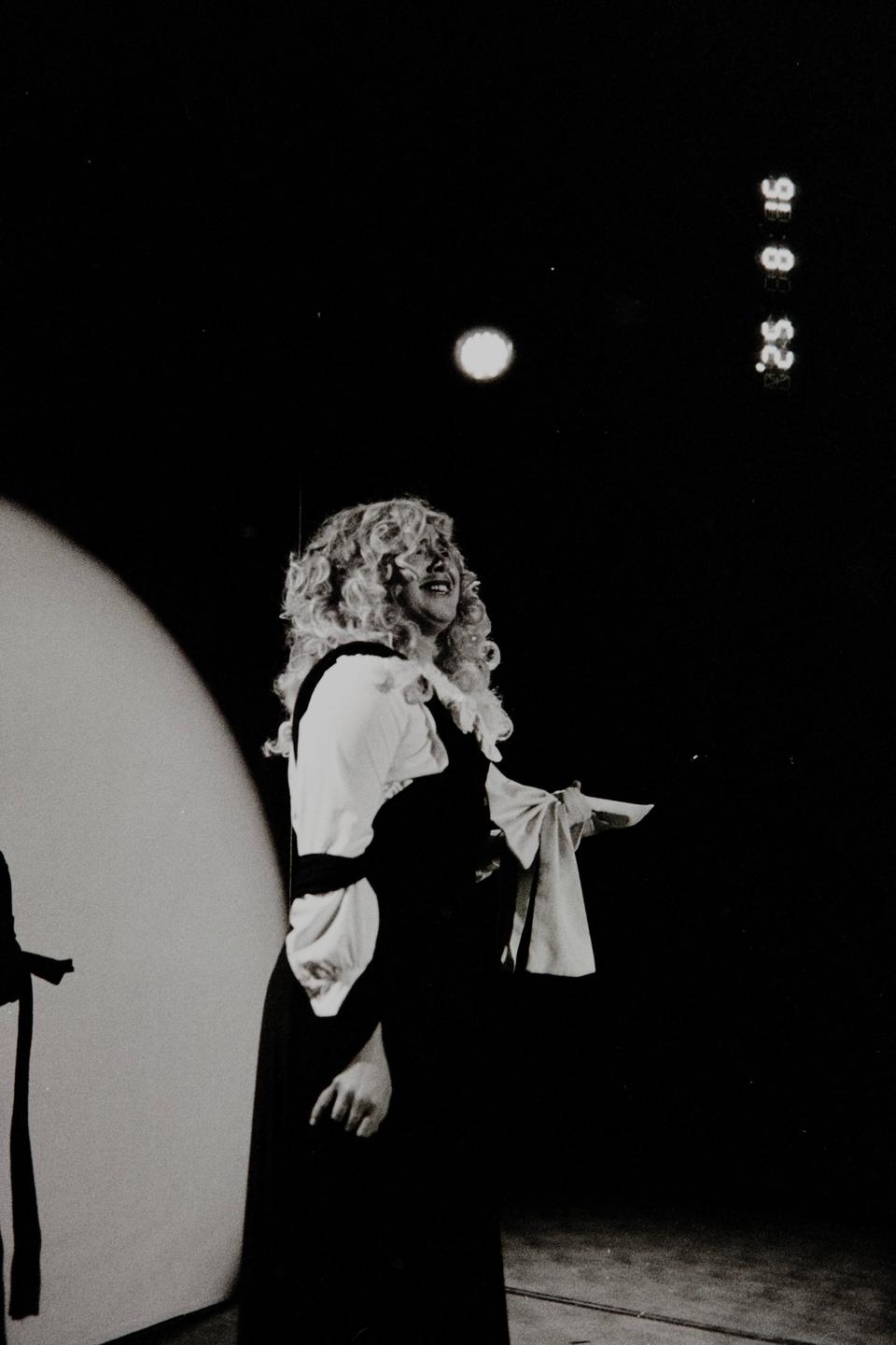 A woman with curly hair and a period costume stands on a stage under a spotlight.