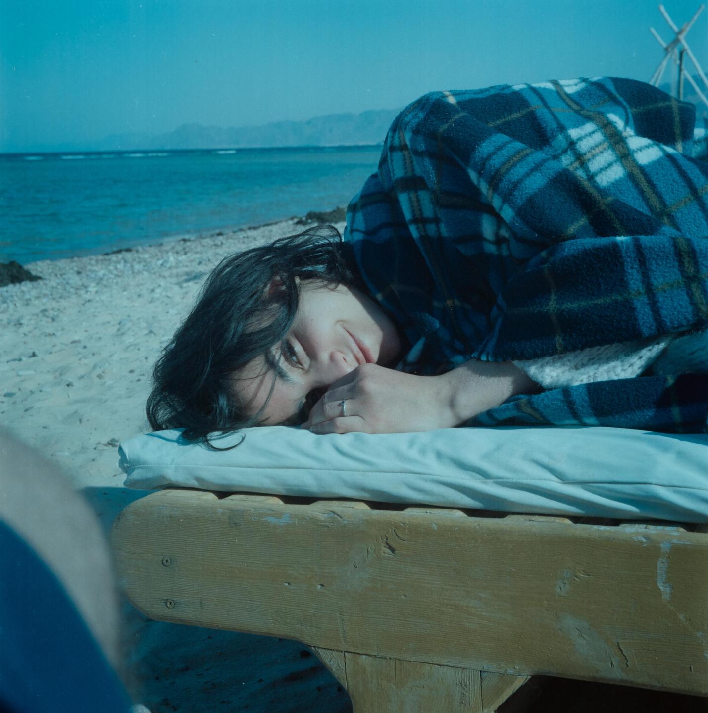 A woman wrapped in a plaid blanket lies on a wooden lounge chair on a sandy beach with the ocean and mountains in the background.