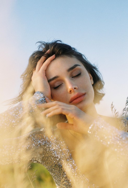 A woman with closed eyes and a sequined top poses with her hand on her head, surrounded by soft-focus foliage.
