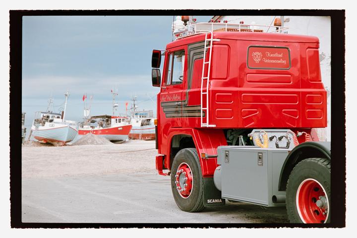 A red truck is parked on a paved surface with several fishing boats visible in the background.
