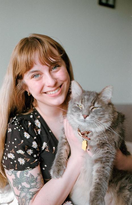 A smiling woman with long hair and a floral dress holds a gray cat with a collar and tag.