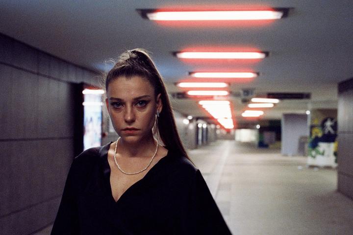 A woman with a ponytail and a pearl necklace stands in a subway station with red neon lights on the ceiling.