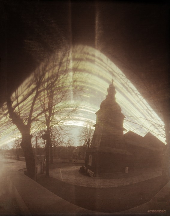 A wooden church with a tall steeple stands next to bare trees under a bright, streaky sky.