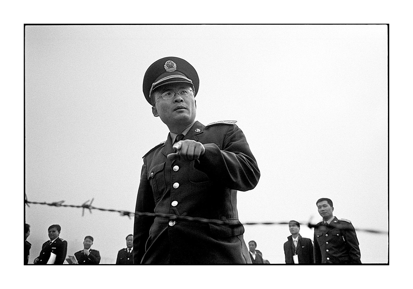 A man in a military uniform and hat points his finger while standing behind barbed wire with other men in the background.