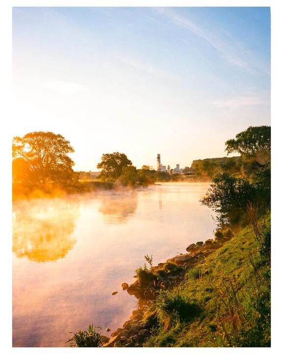 A river with mist rising from its surface flows past a grassy bank and trees under a bright sky with industrial buildings in the distance.