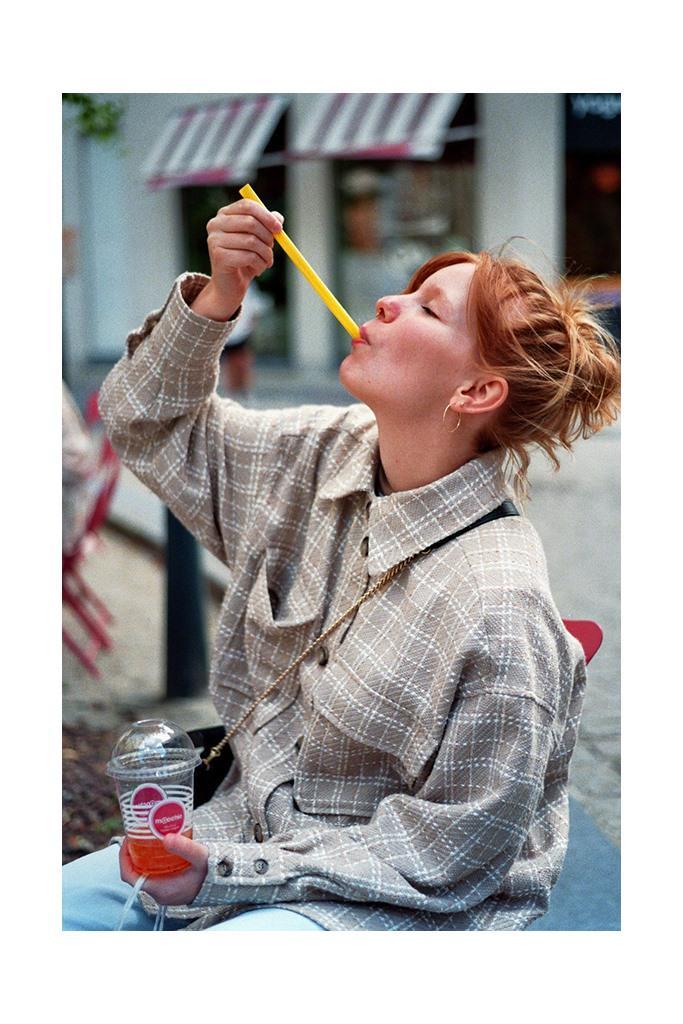 A woman with red hair sips a drink through a yellow straw on a street.