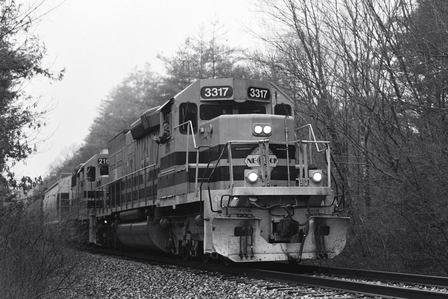 A man waves from the window of a train moving on a track through a forest of bare trees.