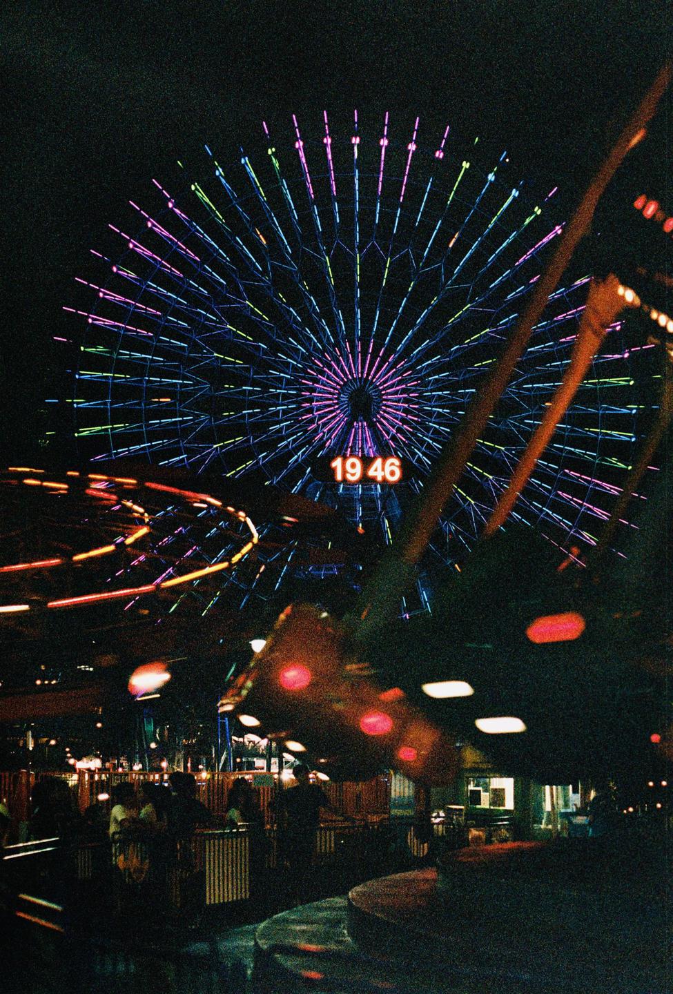 A ferris wheel with colorful lights and a digital clock displaying "19 46" is visible at night.