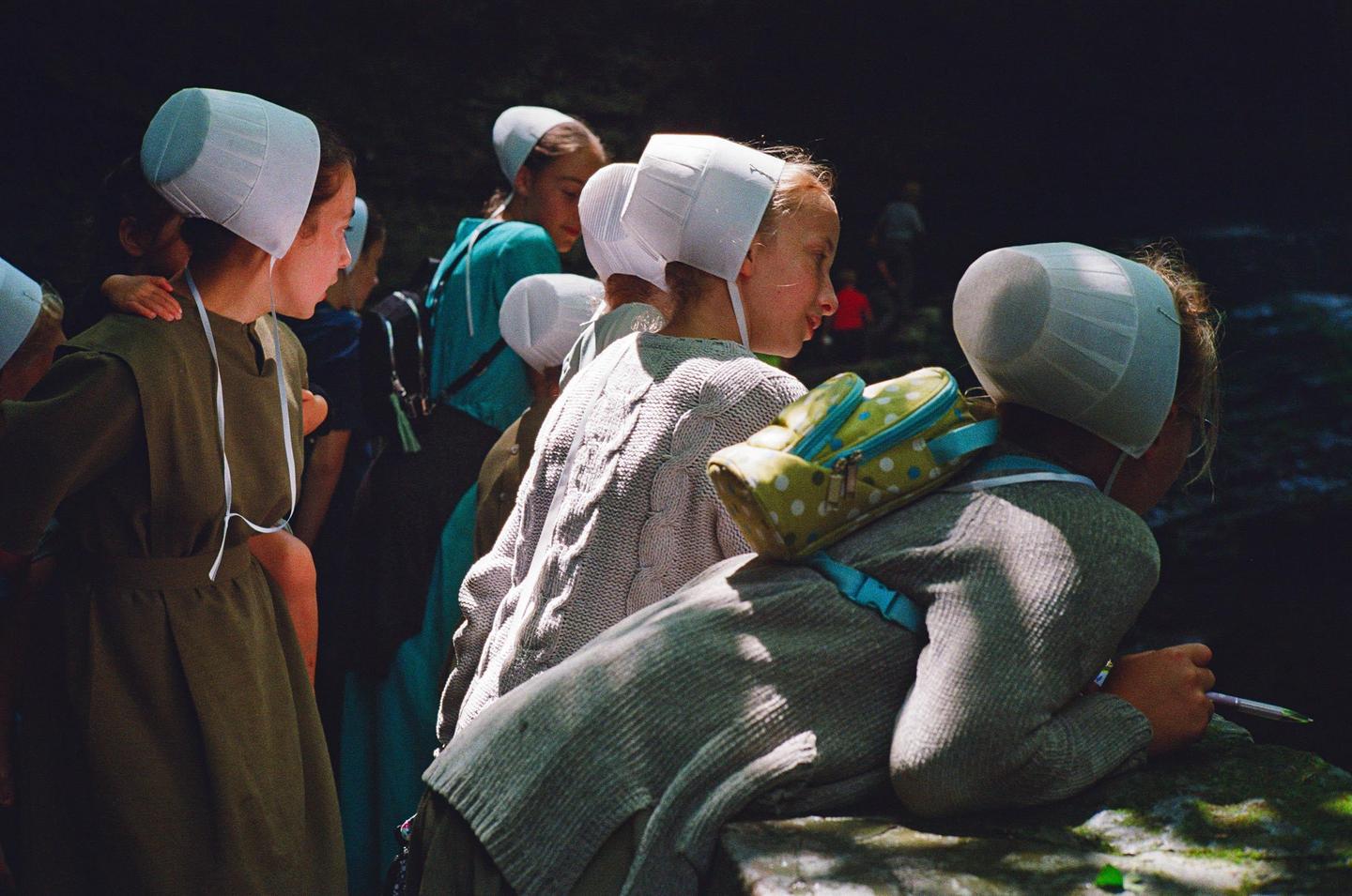A group of girls and women wearing bonnets are gathered in a dark, shaded area, with one girl leaning on a rock and holding a pen.