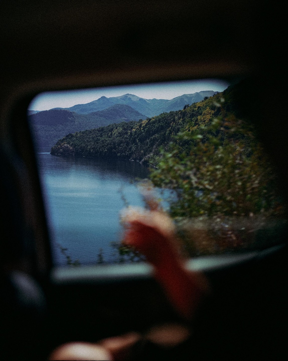 A person's arm is visible in the foreground, with a view of a lake and mountains through a car window.