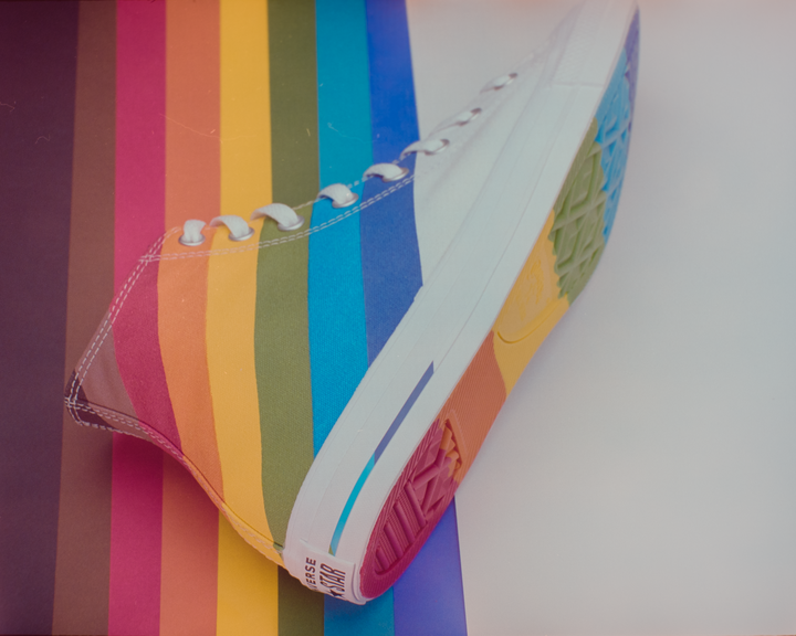 A rainbow-striped high-top sneaker is placed diagonally on a background with vertical rainbow stripes.