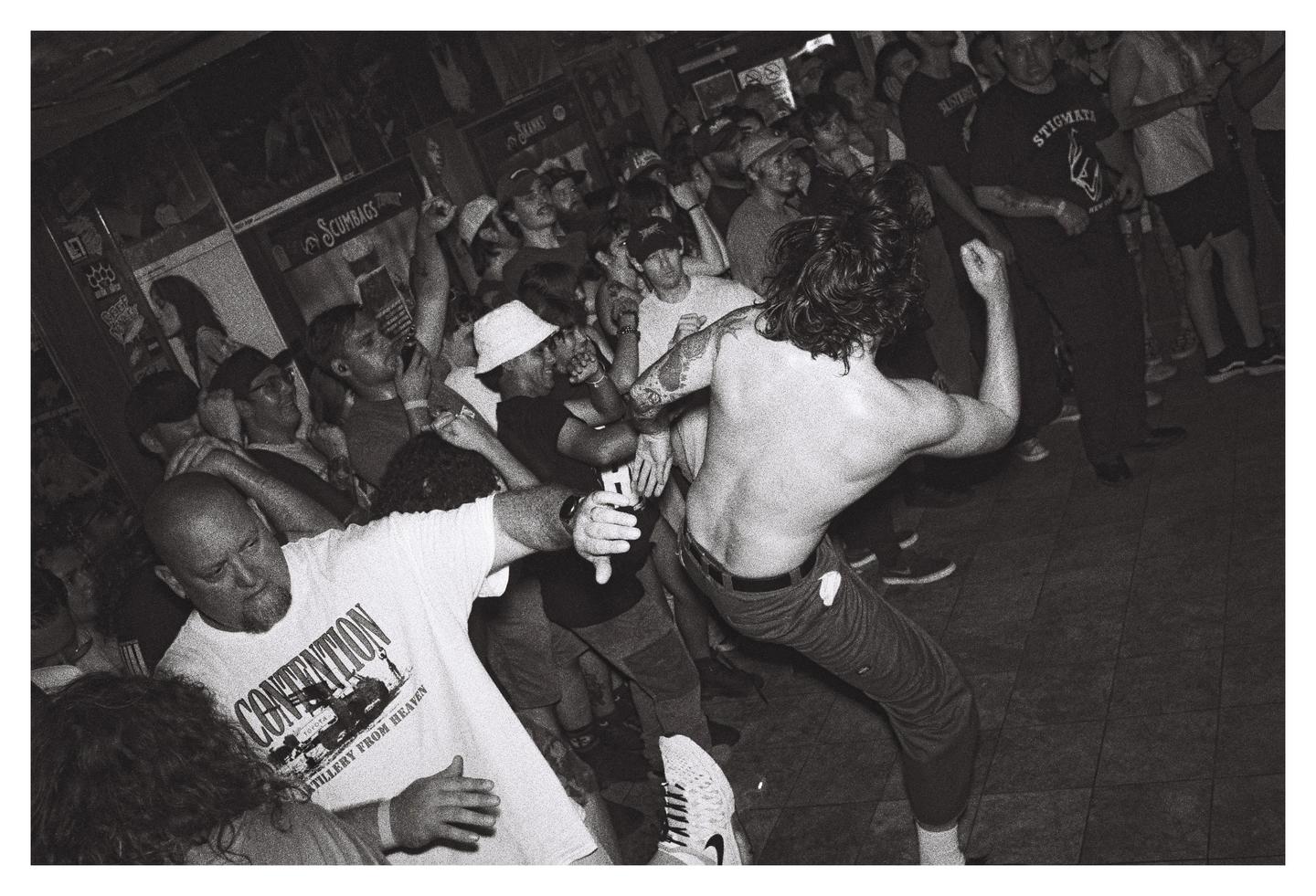 A shirtless man with long hair dances in a crowded room with other men.