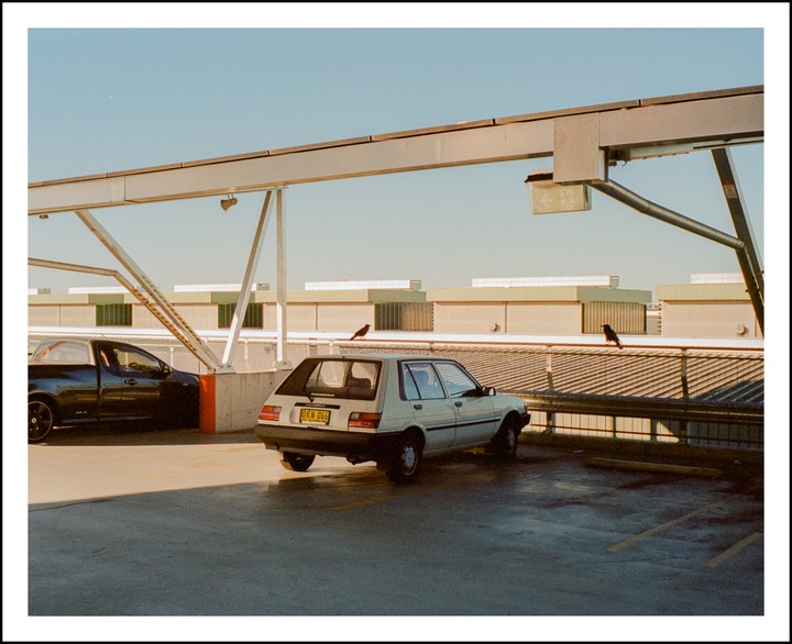 Two cars are parked on the top level of a parking garage with two birds perched on the railing.