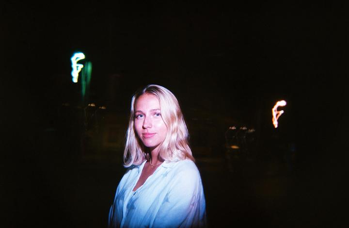 A woman with blonde hair looks at the viewer in a dark setting with blurred lights in the background.