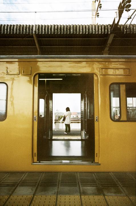 A woman stands on a train platform, visible through the open door of a yellow train car.